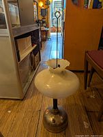 Full view of the oil lamp showing metal base, white glass shade, and wick adjustment rod.