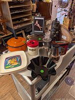 Overview photo of various vintage kitchenware and decor items arranged on a glass display table. Shows enamel pot, ceramic plates, black plastic bowl, framed photo, glass decanter, metal container, and tiki mask.