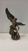 Front view of bronze-finish statuette showing Archangel Michael with sword above and wings spread, standing over Lucifer figure on base.