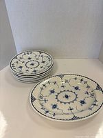 Photo of six round side plates stacked with one oval plate in front; all white porcelain with blue floral pattern.