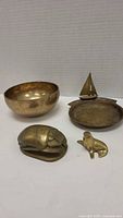 Four brass items arranged on table: bowl, sailboat dish, scarab, and seal figurine.