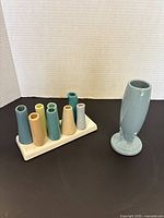 Full view of the vintage blue Fiesta ceramic vase next to the small ceramic vase holder with seven small vases in various pastel colors.