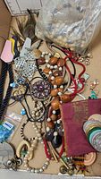 Stacked assortment of various costume jewelry pieces including necklaces, earrings, a peacock feather and plastic tags inside a cardboard box.