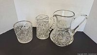 Three cut crystal items arranged side by side on black surface and white background: vase on left, smaller pitcher center, large pitcher right.