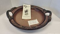 Full view of the large pottery platter showing round shape, glazed surface, decorative rim and handles, with two labels inside the platter.