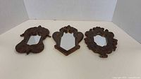Three small handmade mirror frames with dark brown antique finish and detailed vintage style shapes arranged side by side on a white surface.