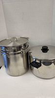 Two stainless steel stock pots seen together, one larger with steam basket insert and one smaller with lid and black handle