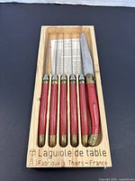 Boxed set showing all six utensils including five forks and one knife with red handles in a wooden box stamped Laguiole de table Fabrique à Thiers - France