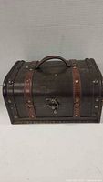 Closed view of a small wooden treasure chest with dark finish and brown leather straps accented with metal studs, featuring a metal latch and leather handle on top.