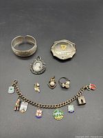 Overview of the lot showing silver bangle, charm bracelet with nine charms, decorative brooch, three rings, and silver compact