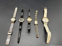 Five vintage ladies watches displayed in a row on a gray surface, mixed band types including metal mesh, leather, and linked metal, varying sizes and shapes.