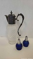 Front view of vintage glass pitcher with metal handle and lid alongside blue glass salt and pepper shakers.