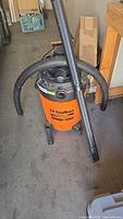 Full view of orange shop vacuum with black hose attached, metal extension tube leaning on vacuum, placed on a concrete floor.