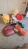 Photo showing automotive items including the Schafer battery charger, Noma booster cables in red bag, trouble light with orange cord, trailer hitch ball, fuel container, yellow cloths, sponge in plastic, and yellow caddy.