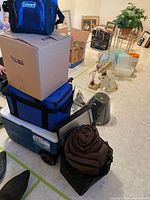 Several coolers stacked including a large hard-shell wheeled cooler and multiple soft cooler bags with Coleman branding. Rolled brown sleeping bag in black carry bag placed in front. Various loose household items in background.