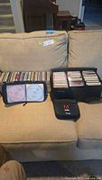 Photo showing an open cassette case filled with music cassettes and a separate case holding CDs on a beige couch