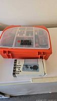 Orange and black Black & Decker plastic case with transparent lid showing compartments full of drill bits and screwdriver bits, with Black & Decker logo on lid.