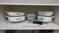 Five white Corningware dishes with cornflower blue floral pattern, varying in size with lids placed on or beside them. Two metal racks and one black detachable handle placed on the shelf with the dishes.