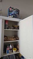 White cabinet with shelves containing assorted crafting wood pieces on upper two shelves and adhesives, glues, sprays and small containers on bottom shelf. Trex handheld vacuum in original box on top of cabinet.
