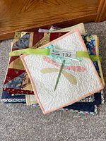 Stack of folded handmade quilts in various patchwork designs and colors with a smaller quilted placemat featuring a dragonfly applique in front.