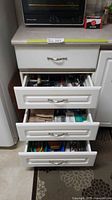 Three white drawers filled with various kitchen tools and accessories, view of the closed drawers with one drawer open showing contents.