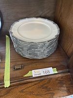 Stack of twelve white bone china dinner plates with scalloped edges and decorative blue and silver floral pattern around the rim, placed on a wooden shelf.