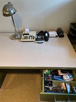 Full desk view with office lamp, adding machine, tape dispenser, stapler, calculator, and partial view of open desk drawer filled with various office supplies.