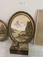 Full view of oval oil painting in vintage gold-tone ornate frame showing mountain and cabin scene, with keepsake box beneath