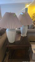 Pair of table lamps with white frosted glass bases and beige pleated fabric shades positioned on a wooden side table in a living room.