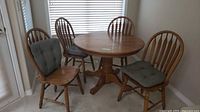 Round pedestal solid oak kitchen table with 4 matching chairs situated in a corner near windows.