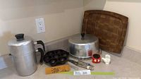 Photo of vintage kitchen items including aluminum coffee percolator, cast iron pan, Mickey Mouse shakers, aluminum cake cover, wooden cutting board, metal tongs, and measuring stick on kitchen counter.