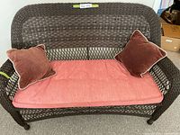 Front view of dark brown wicker bench with pink seat cushion and two brown velvet pillows.