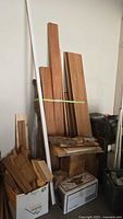 Two images showing various boards of hardwood lumber leaning against a wall with boxes at base containing additional wood and shingles.