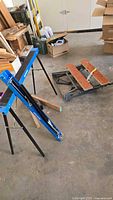 Full view of blue Makita folding saw horses with black legs and folded Workmate bench with orange wooden planks, alongside two steel saw horse brackets.