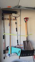 Photo showing various yard tools hung on wall including wide push broom, smaller broom, shovels, and dustpan.