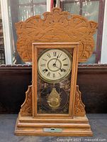 Front view of the oak gingerbread clock showing carved ornate wooden design, Roman numeral clock face, pendulum behind etched glass.