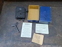 Complete set including Meccano motor, winding key, black cylinder part, original box, and instruction sheets on wooden table.