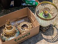 Two early table lamps shown outdoors, one partially assembled with ornate brass and crystal details, other disassembled in a cardboard box