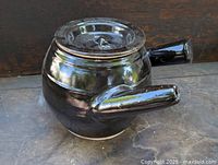Brown glazed pottery crock with lid and two handles set on a stone surface, side angle view showing one handle and lid.