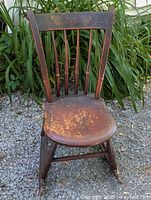 Front view of early 1800s pine wooden rocking chair showing original dark finish with wear and carved backrest details. Taken outdoors with greenery background.