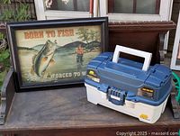 Framed fishing art print and Plano tackle box shown together on a surface