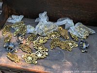 Lot of vintage brass drop handles and ornate backplates, some in plastic bags, arranged on wooden surface.