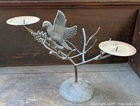 Front view of metal bird sculpture candle holder on wooden surface, showing bird and two candle plates on branch