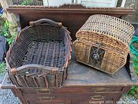 Photo of two wicker baskets on a wooden surface showing the large dark brown basket and the smaller one with a hinged lid and latch