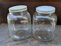 Pair of vintage clear glass square-shaped storage jars with white metal screw-on lids, placed side by side on a wooden surface in natural lighting.