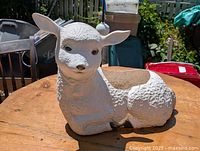 Front-right angled view of white lamb-shaped plastic flower pot on wooden surface outside showing facial details and hollow planting area.