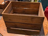 Rectangular open pine wood box with handles, placed on wooden surface under daylight, showing front and side views with visible wood grain and slight wear.