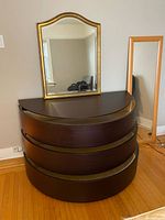 Photo showing semi-circle dark brown wooden bureau with three flush drawers, with two mirrors resting on the top: a gold framed decorative mirror and a taller, light wood framed full-length mirror
