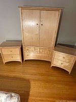 Front view of the light wood armoire with closed doors and matching bedside tables on either side, showing two wide drawers on the armoire and two drawers on each bedside table.