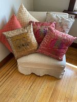 Photo of a light beige slip-covered casual chair with six assorted throw pillows, including red patterned, gold with patterns, pale damask, and orange pillows, placed on a wooden floor next to a window.